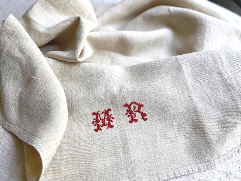 May include: Cream-colored linen cloth with red embroidered initials "MR". The fabric is folded, showing texture and slight variations in color. The embroidery is in a decorative, stylized font.