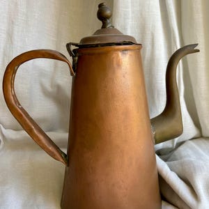 Könnte beinhalten: Eine Vintage-Kaffeekanne aus Kupfer mit einem langen, gebogenen Ausguss und einem abgerundeten Griff. Die Kanne hat einen konischen Körper und einen kleinen, runden Deckel mit einem Knopf. Griff und Ausguss haben eine andere Farbe als der Körper.