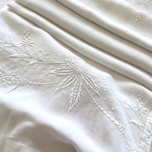 May include: White linen bedding with detailed embroidered floral and leaf patterns. The embroidery features a palm leaf design and delicate dotted accents. The bedding is folded, showcasing the intricate craftsmanship and texture.