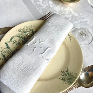 May include: A white, rolled-up linen napkin with embroidered initials, resting on a vintage plate with green floral accents. Silverware and a patterned tablecloth complete the elegant table setting.