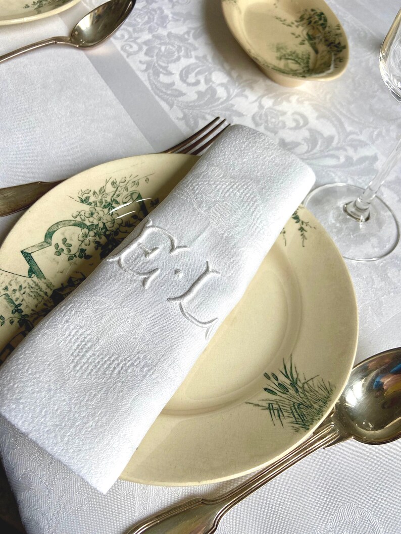 May include: A white, rolled-up linen napkin with embroidered initials, resting on a vintage plate with green floral accents. Silverware and a patterned tablecloth complete the elegant table setting.