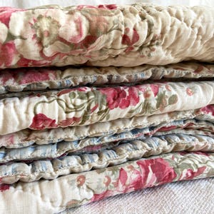Pode incluir: Uma manta acolchoada dobrada com um padrão floral em tons de rosa, vermelho e verde sobre um fundo branco sujo. O verso do tecido apresenta um padrão de riscas finas em azul claro e branco visível ao longo das bordas dobradas.