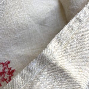 May include: Close-up of a beige linen fabric with a visible weave. The fabric has a slightly textured appearance and a natural, off-white color. A red design is visible in the corner of the fabric.