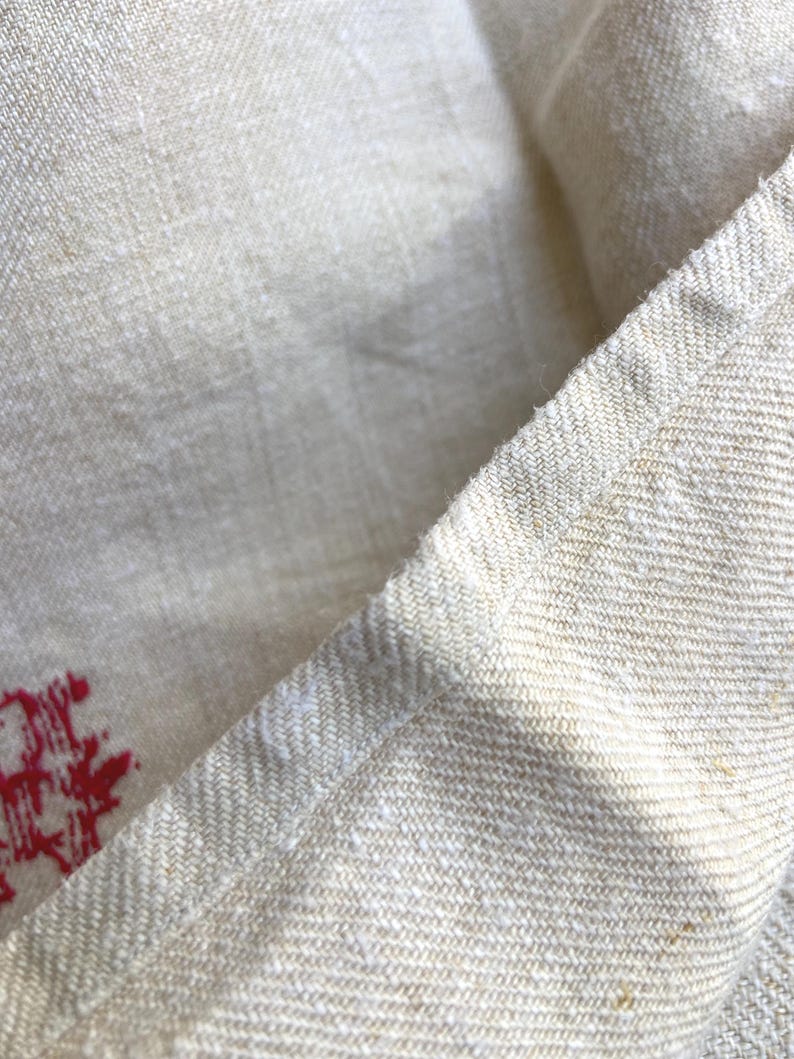 May include: Close-up of a beige linen fabric with a visible weave. The fabric has a slightly textured appearance and a natural, off-white color. A red design is visible in the corner of the fabric.