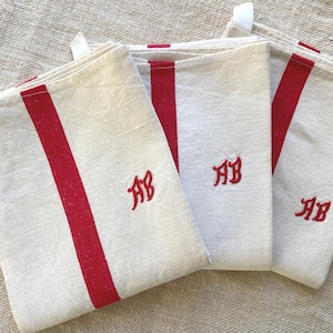 May include: Three rectangular off-white kitchen towels featuring a vertical red stripe running down the center and the embroidered red monogram letters AB. Each towel includes a small white fabric loop attached to the top corner for hanging.