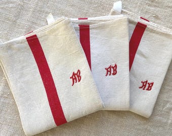 May include: Three rectangular off-white kitchen towels featuring a vertical red stripe running down the center and the embroidered red monogram letters AB. Each towel includes a small white fabric loop attached to the top corner for hanging.