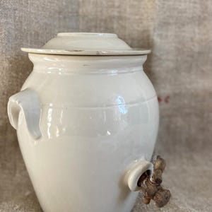 May include: White ceramic crock with a lid and a wooden spigot. The crock has a rounded body and a wide mouth. The spigot is attached to the side of the crock and has a wooden handle.