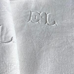 May include: White linen napkins with an embroidered monogram. The napkins are folded and stacked, with one rolled up, revealing the intricate texture and design. The monogram features the letters "DL".