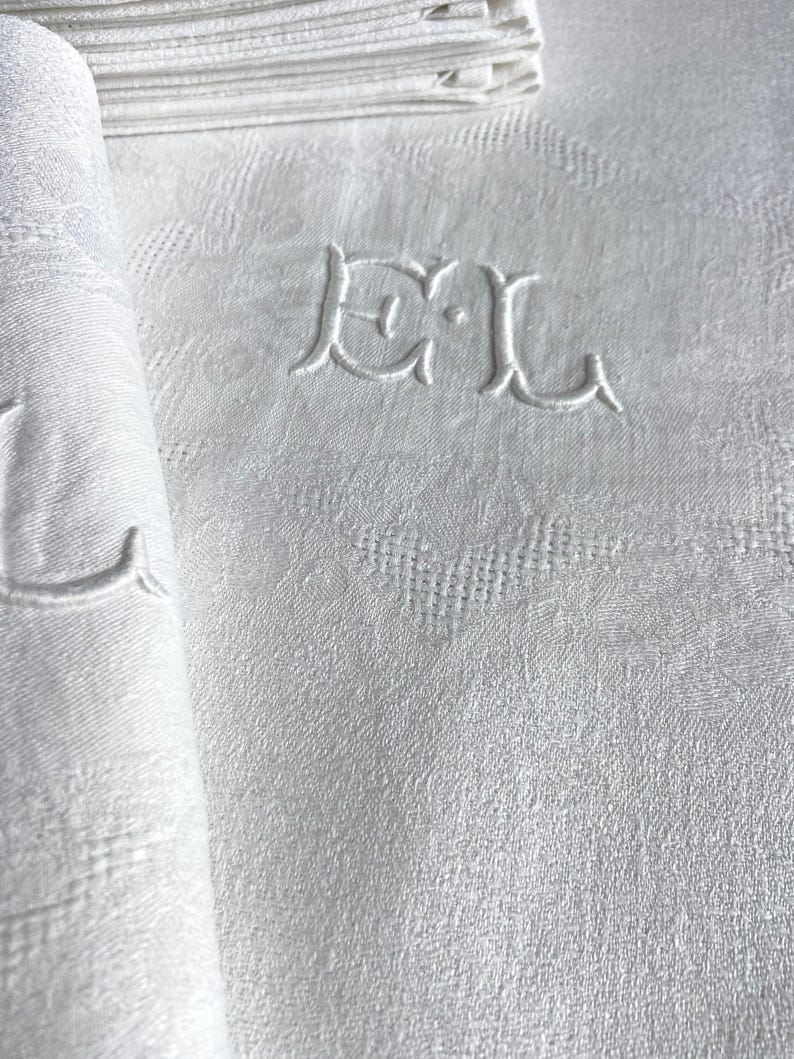 May include: White linen napkins with an embroidered monogram. The napkins are folded and stacked, with one rolled up, revealing the intricate texture and design. The monogram features the letters "DL".