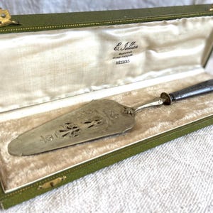 May include: An antique silver cake server with a decorative handle and ornate blade, presented in a green velvet-lined box. The box has gold-colored clasps and hinges. The server is engraved with a floral design.