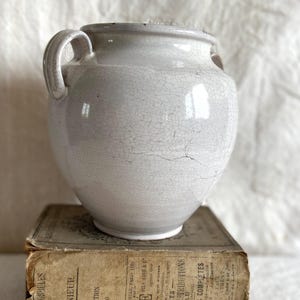 Peut inclure: Un pot en céramique blanc cassé vieilli, avec deux anses et un effet de glaçure craquelée. Il repose sur un livre ancien avec du texte décoloré. L'ensemble évoque un style rustique et un savoir-faire artisanal. Mesures en cm.