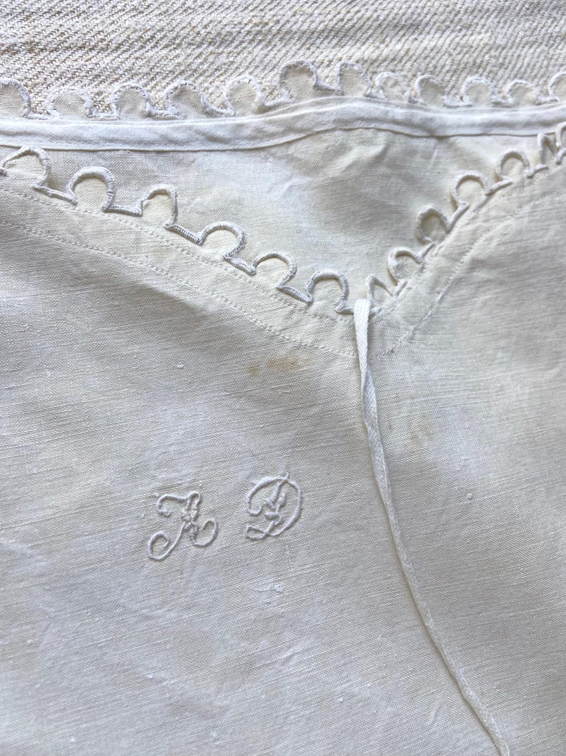 Puede incluir: Un tejido de lino en color blanco roto que presenta un borde festoneado a lo largo de la parte superior. La tela muestra un monograma bordado con las letras A y D en letra cursiva. El material tiene una textura tejida visible y una ligera decoloraci&oacute;n.