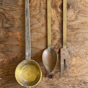 May include: A set of three vintage kitchen utensils featuring a large round brass ladle with a long handle, a smaller oval-shaped copper spoon with a long brass handle, and a two-pronged brass meat fork with a long handle and metal rivets.