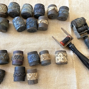 May include: An assortment of vintage, cylindrical wooden stamps with carved designs in various colors, including blue, beige, and brown. Two roller stamps with black handles are also present. The stamps are arranged on a neutral-toned surface.