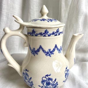 May include: Cream ceramic teapot with blue floral and leaf patterns. The teapot features a curved handle, a spout, and a lid with a finial. The blue design is repeated around the top and bottom of the teapot.