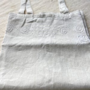 May include: A white linen tote bag with two straps and a decorative embroidered design. The embroidery features a repeating pattern of circles and lines.
