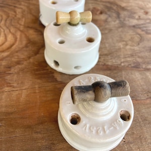 Op de afbeelding: Drie ronde elektrische schakelaars van wit keramiek met een glanzende afwerking. Elke schakelaar heeft een centrale draaibare houten tuimelschakelaar. De basis van de voorste schakelaar is voorzien van de tekst 6A-125V en 10A-250V.