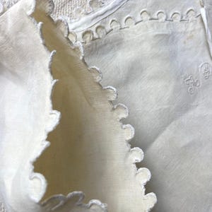 Puede incluir: Una pieza textil de color blanco roto con bordes festoneados y bordados decorativos. La tela muestra una textura tejida e incluye un peque&ntilde;o detalle de monograma bordado que consiste en las letras L y D en la superficie del material.