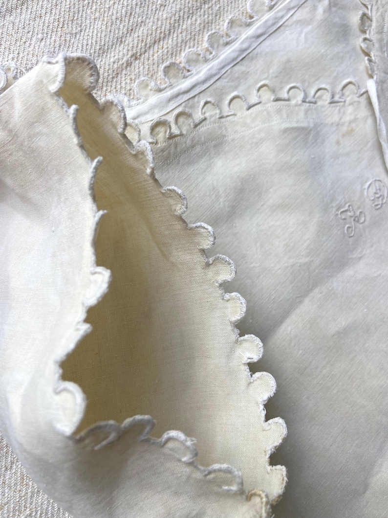 Puede incluir: Una pieza textil de color blanco roto con bordes festoneados y bordados decorativos. La tela muestra una textura tejida e incluye un peque&ntilde;o detalle de monograma bordado que consiste en las letras L y D en la superficie del material.