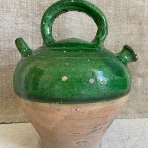 May include: A green glazed ceramic pitcher with a rounded body and a single handle. The pitcher has a spout on each side and a brown unglazed base.