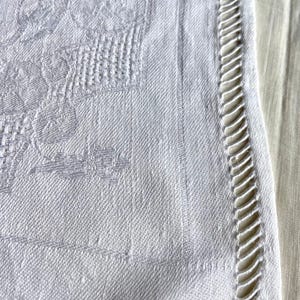 May include: Close-up of a white linen tablecloth with a floral pattern and a decorative, cutwork edge. The fabric has a subtle texture and a vintage aesthetic, suitable for a dining table or decorative use.