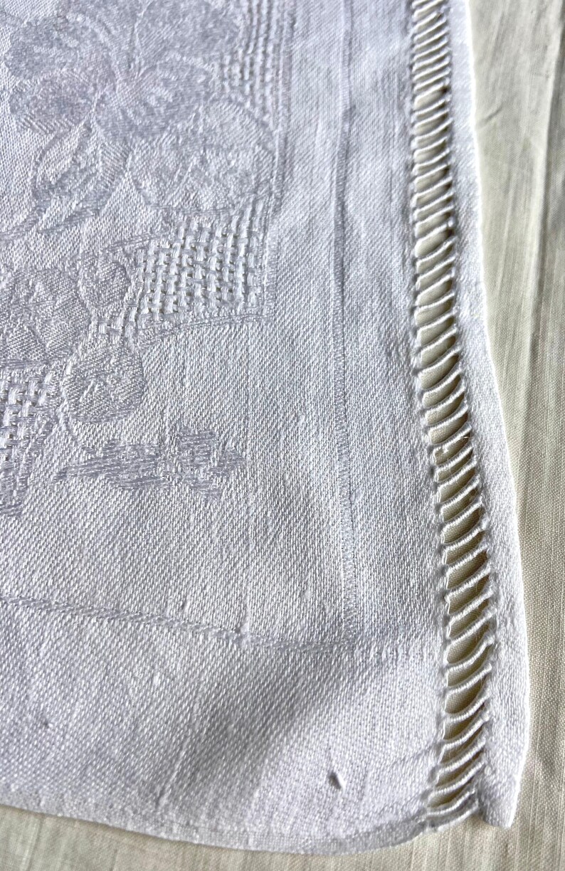 May include: Close-up of a white linen tablecloth with a floral pattern and a decorative, cutwork edge. The fabric has a subtle texture and a vintage aesthetic, suitable for a dining table or decorative use.