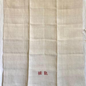 May include: A folded, off-white linen cloth with three vertical panels. The cloth has a subtle texture and is embroidered with the red letters "MR" in a decorative font. The cloth appears to be vintage or antique.
