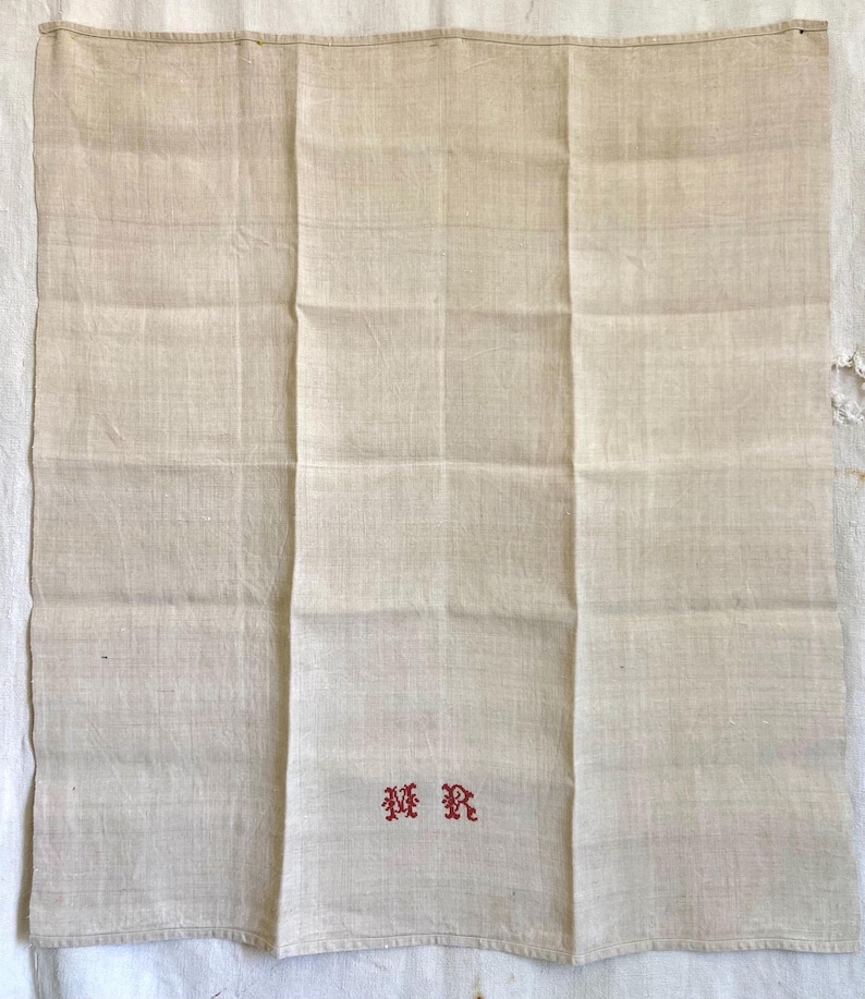 May include: A folded, off-white linen cloth with three vertical panels. The cloth has a subtle texture and is embroidered with the red letters "MR" in a decorative font. The cloth appears to be vintage or antique.