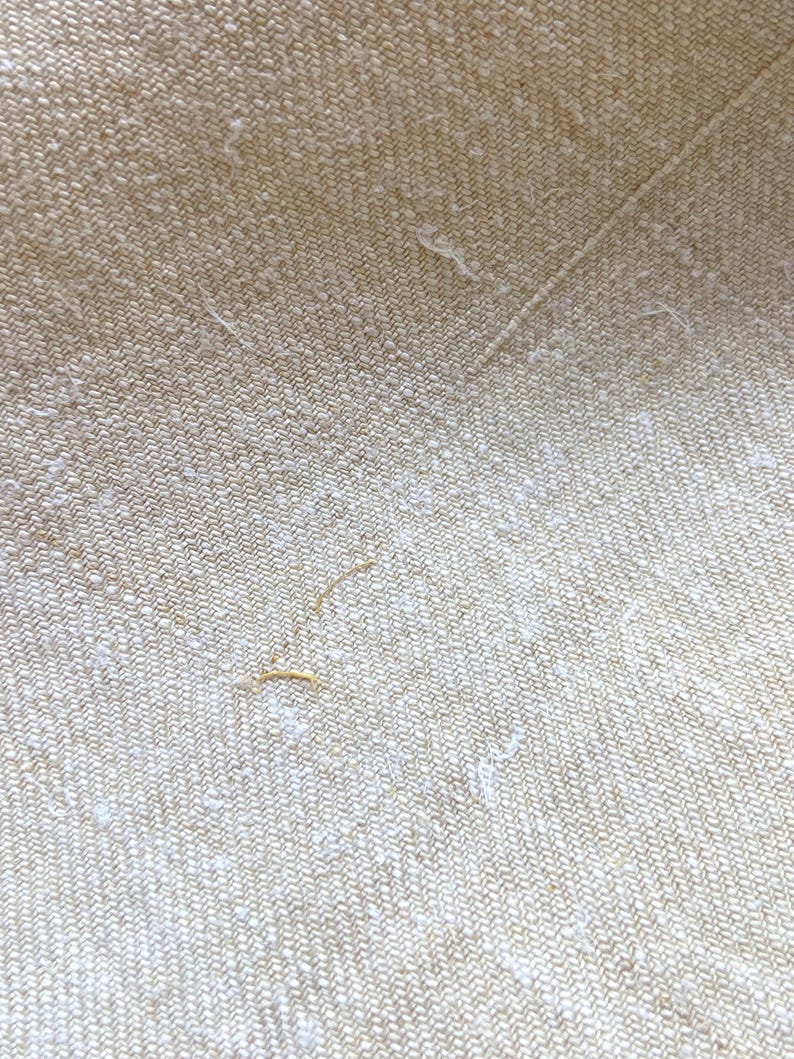 May include: Close-up of a beige fabric with a subtle herringbone weave. The texture is slightly rough, with small, irregular white flecks and a few loose threads. The fabric has a natural, organic appearance.
