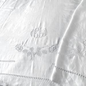 Pode incluir: Um têxtil branco com intrincados bordados florais em branco sobre branco, motivos de rosas e um monograma central. O tecido inclui uma bainha decorativa aberta ao longo das bordas, criando um padrão detalhado na superfície branca.