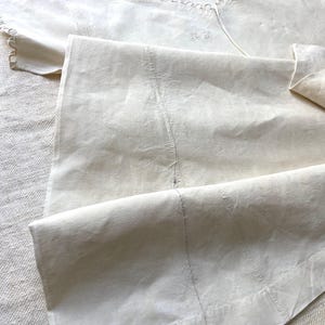Puede incluir: Una prenda de color crema, blanco roto, con un escote ancho adornado con bordes festoneados decorativos y bordados calados. La tela muestra costuras verticales visibles y un detalle de monograma sutil cosido cerca del cuello.