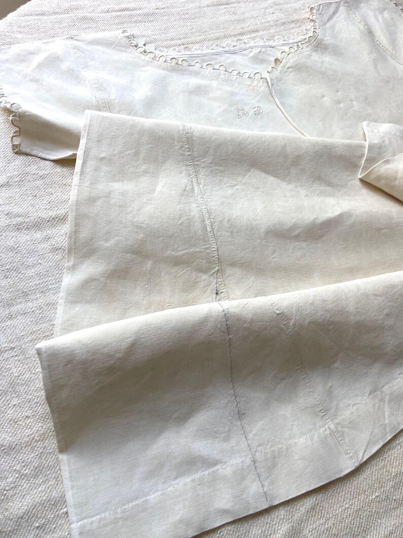 Puede incluir: Una prenda de color crema, blanco roto, con un escote ancho adornado con bordes festoneados decorativos y bordados calados. La tela muestra costuras verticales visibles y un detalle de monograma sutil cosido cerca del cuello.