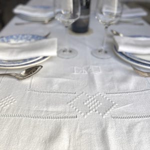 Pode incluir: Uma toalha de mesa de linho branco com padrões geométricos bordados está montada para um jantar formal. A mesa está posta com pratos, talheres, taças de vinho e guardanapos brancos dobrados. Uma garrafa de vinho está no fundo.