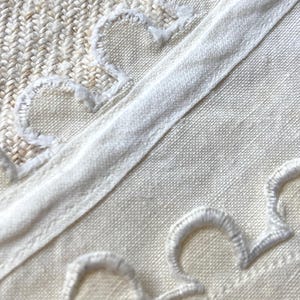 Puede incluir: Un tejido de color crema que presenta una banda de tela recta y lisa que atraviesa diagonalmente el centro. Los bordes de la banda est&aacute;n adornados con bordados festoneados blancos, creando un patr&oacute;n decorativo de semic&iacute;rculos.