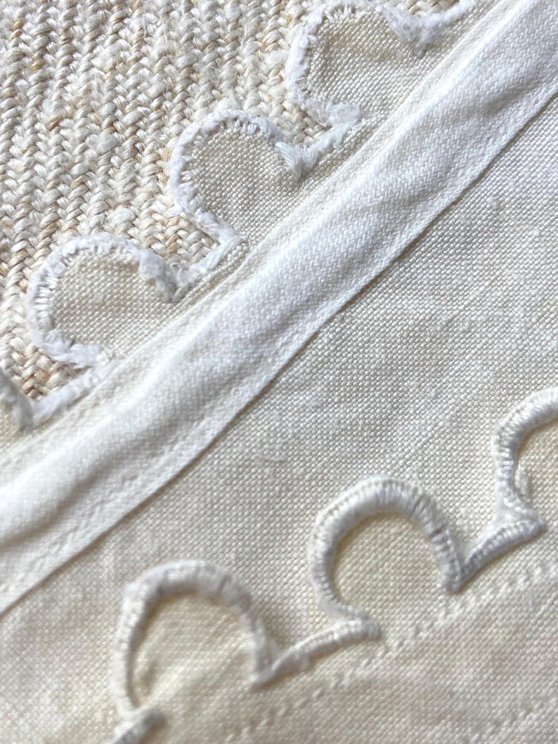 Puede incluir: Un tejido de color crema que presenta una banda de tela recta y lisa que atraviesa diagonalmente el centro. Los bordes de la banda est&aacute;n adornados con bordados festoneados blancos, creando un patr&oacute;n decorativo de semic&iacute;rculos.