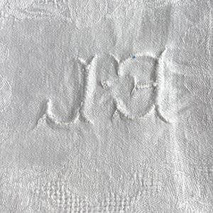 May include: Close-up of a white textile with a floral pattern. The fabric features embroidered initials "J.J" in a delicate, cursive font. The background is a detailed, textured weave, suggesting a vintage or heirloom piece.