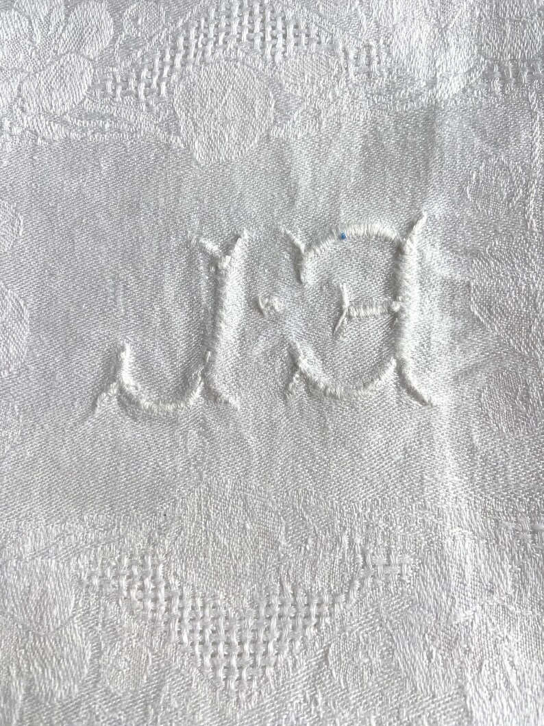 May include: Close-up of a white textile with a floral pattern. The fabric features embroidered initials "J.J" in a delicate, cursive font. The background is a detailed, textured weave, suggesting a vintage or heirloom piece.
