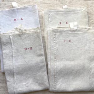 May include: A collection of white linen napkins, each embroidered with initials in a reddish-pink thread. The napkins are folded and stacked, with a small loop at the top. The fabric has a slightly textured appearance.