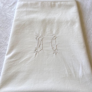 Peut inclure: Un tissu de lin plié, de couleur blanc cassé, avec les initiales "MC" brodées dans un motif floral délicat. La broderie est réalisée avec un fil blanc cassé assorti, créant un contraste subtil et élégant. L'article semble être vintage ou ancien.