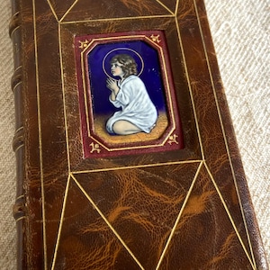 Könnte beinhalten: Ein Vintage-Buch in braunem Leder mit goldenen Akzenten. Das Cover zeigt einen rechteckigen Einsatz mit einem gemalten Bild einer betenden Figur vor blauem Hintergrund. Die Kanten des Buches sind abgenutzt, was auf das Alter hindeutet.