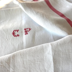 May include: A white linen kitchen towel featuring a red embroidered monogram with the letters C and P. The fabric has a visible woven texture and includes a single vertical red stripe running along the side of the folded cloth.