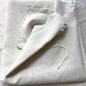 Puede incluir: Una pieza textil de color blanco roto plegada con un borde festoneado y recortes circulares. La tela tiene un aspecto liso y mate, e incluye un cord&oacute;n fino a juego que se extiende desde la esquina de la secci&oacute;n plegada.