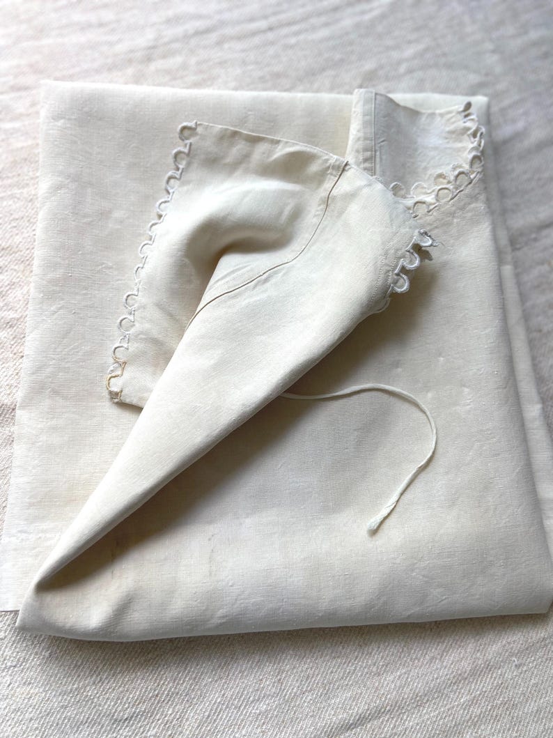 Puede incluir: Una pieza textil de color blanco roto plegada con un borde festoneado y recortes circulares. La tela tiene un aspecto liso y mate, e incluye un cord&oacute;n fino a juego que se extiende desde la esquina de la secci&oacute;n plegada.