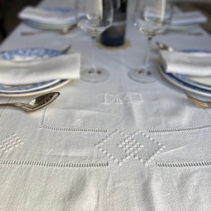 May include: White linen tablecloth with embroidered details and a monogrammed "DM" in the centre. Two wine glasses and plates are set on the table.