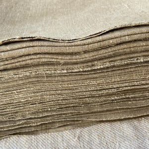 May include: A stack of natural linen fabric, folded neatly. The fabric is a light beige color and has a slightly rough texture.