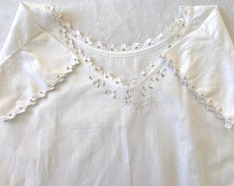 Op de afbeelding: Een wit katoenen kledingstuk met een wijde halslijn en korte mouwen, beide afgewerkt met ingewikkeld wit broderie anglaise. De stof is glad en lichtgewicht, met een delicaat bloemmotief gestikt in de kanten rand.