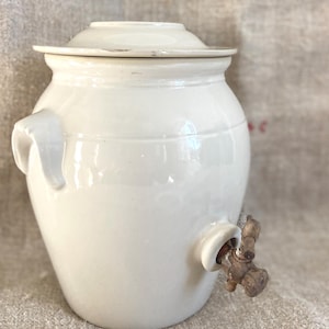 May include: A cream-coloured ceramic crock with a matching lid, featuring two side handles and a metallic spigot attached to the lower front section. The vessel has a smooth, glossy finish and a rounded body shape, designed for liquid storage.