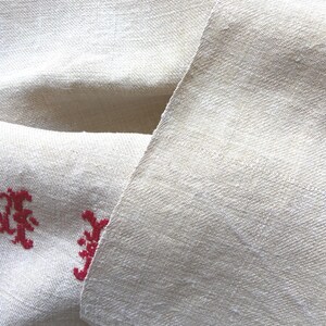 May include: Close-up of a folded, off-white linen fabric with a subtle woven texture. The fabric has a natural, slightly wrinkled appearance. Red embroidered floral design is visible on the lower left corner of the fabric. The fabric is likely used for home decor.