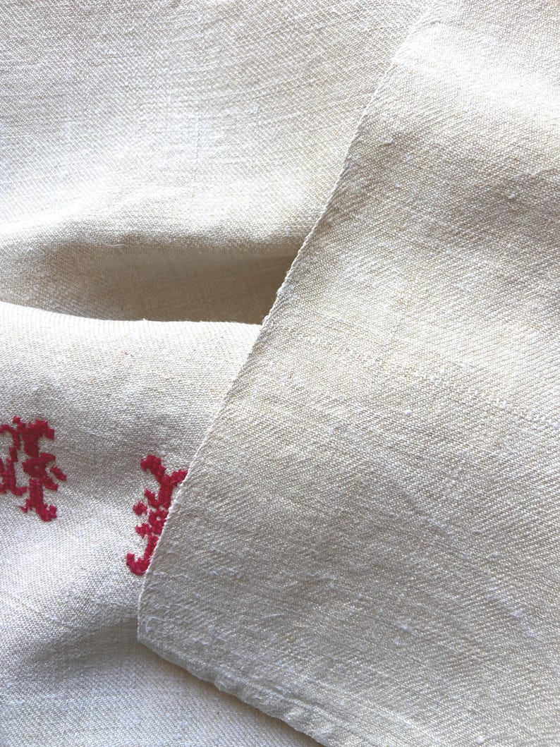 May include: Close-up of a folded, off-white linen fabric with a subtle woven texture. The fabric has a natural, slightly wrinkled appearance. Red embroidered floral design is visible on the lower left corner of the fabric. The fabric is likely used for home decor.