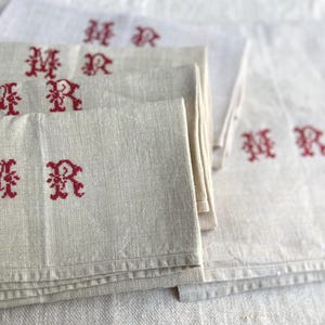 May include: Several off-white linen napkins with red embroidered initials "MR". The napkins are folded and stacked, with some visible in the background. The fabric has a textured appearance.